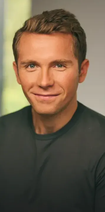 Portrait of a young man with short brown hair and blue eyes smiling while wearing a dark shirt.